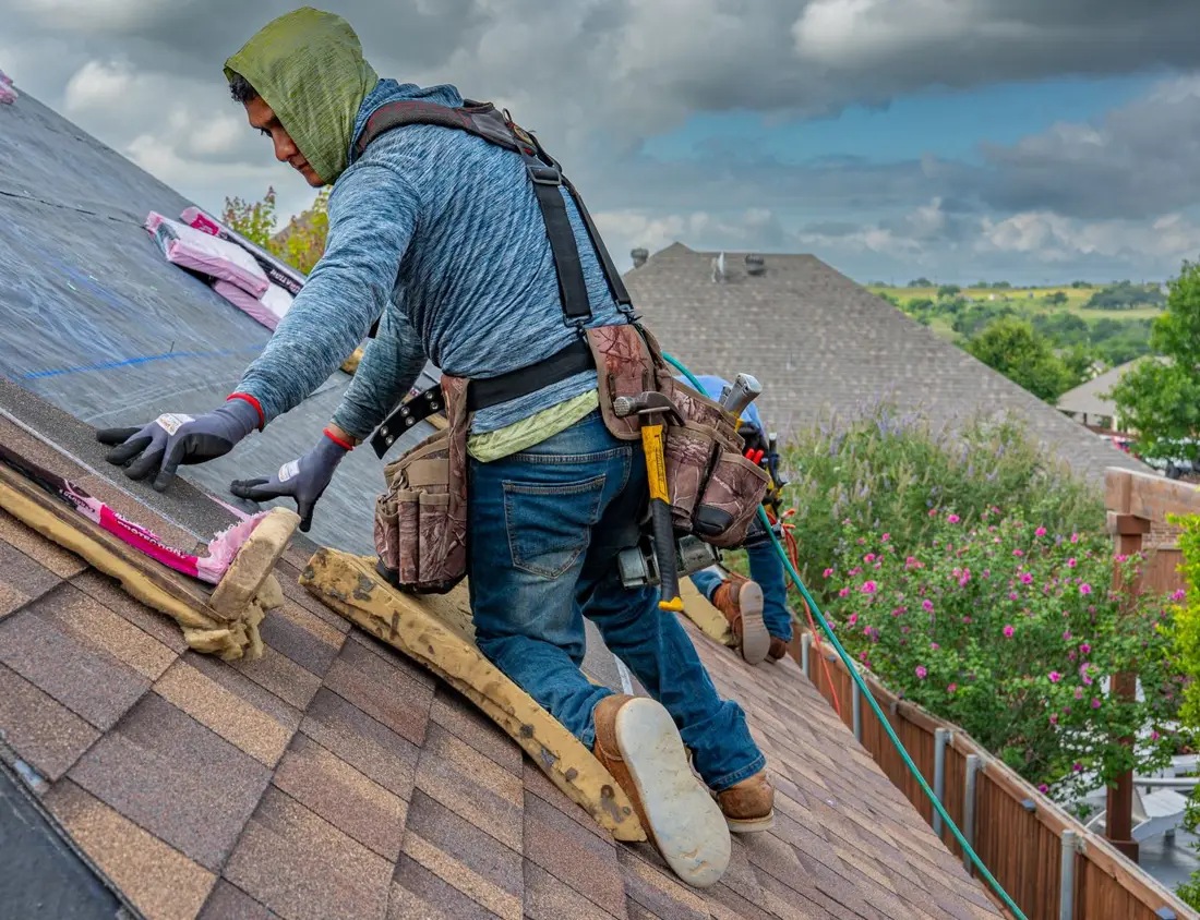 A Ridgewell roofer in a safety harness laying architectural shingles under a cloudy Oklahoma sky